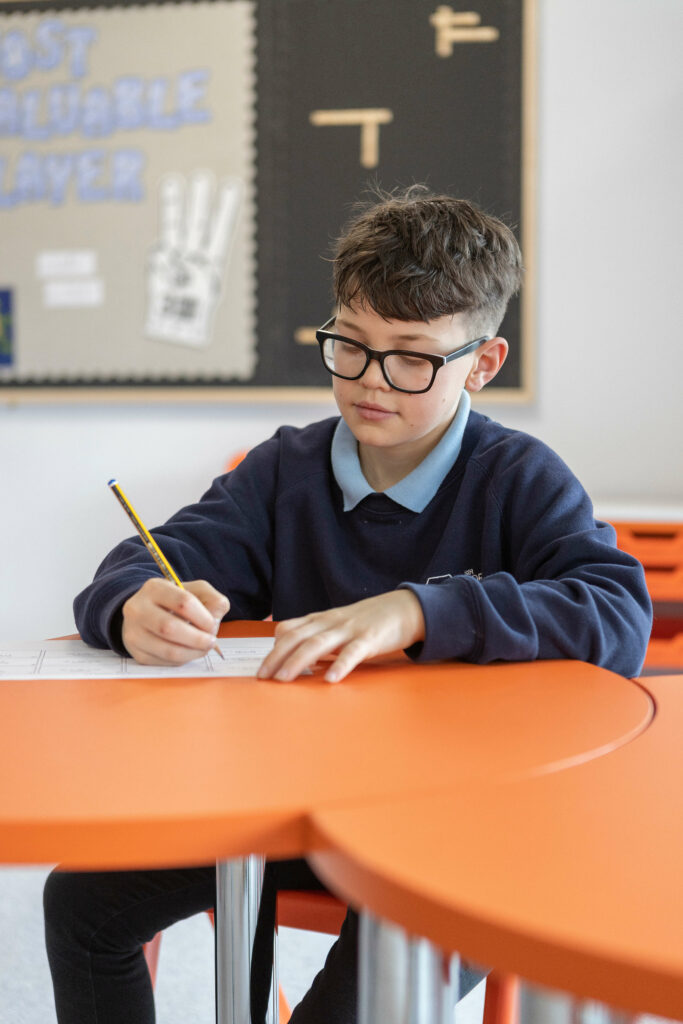 Photo of a pupil sitting at an orange desk and writing in a classroom